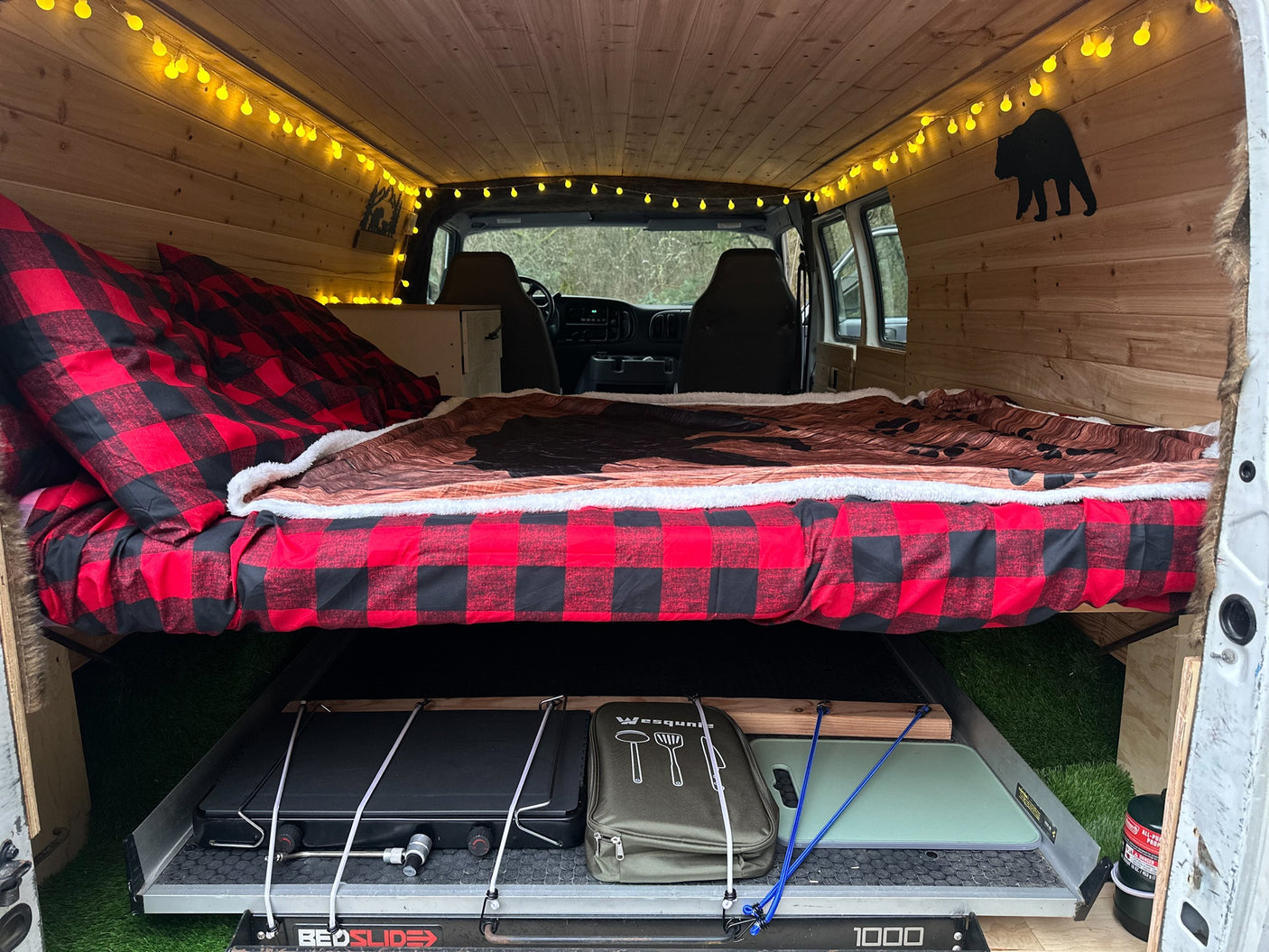 Inside an RV rental: a wood-paneled camper with a cozy bed, string lights, and a pull-out kitchen area below.