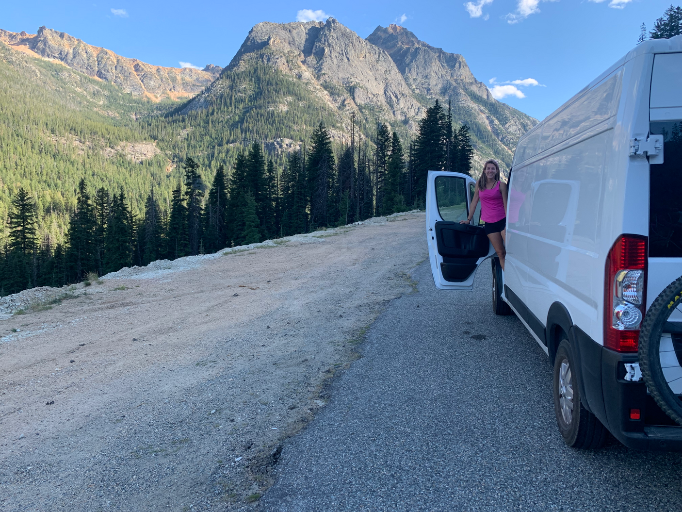 RV rental adventure: Woman smiles by her white camper van, parked amidst stunning mountains & lush forests under blue skies.