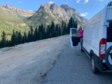 RV rental adventure: Woman smiles by her white camper van, parked amidst stunning mountains & lush forests under blue skies.