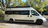 Silver Winnebago Travato camper van, an ideal RV rental, parked on gravel amidst green trees.