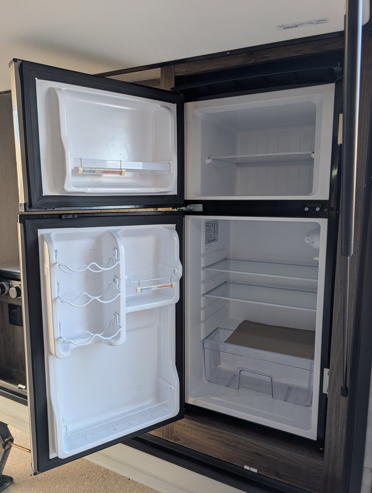 Open RV rental refrigerator/freezer, empty and clean, with shelves, door storage, a thermometer, and wine rack visible.