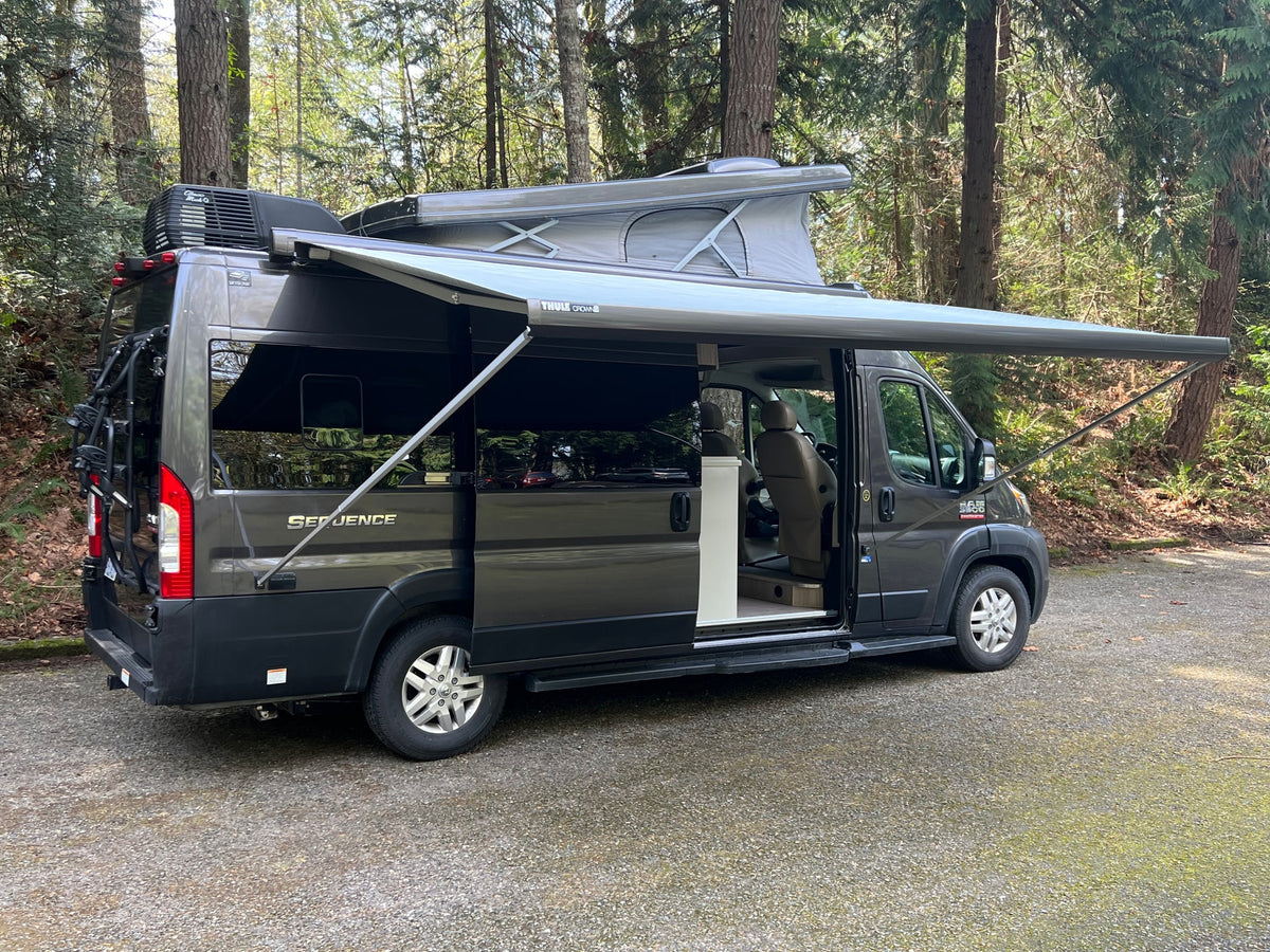 Dark grey pop-top camper van rv rental with extended awning in a wooded setting, perfect for outdoor travel.