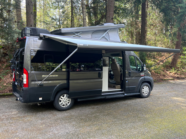 Dark grey pop-top camper van rv rental with extended awning in a wooded setting, perfect for outdoor travel.
