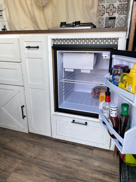 Inside this RV rental: a compact kitchen with an open mini-fridge, small stovetop, and white cabinets. Ready for your adventure!