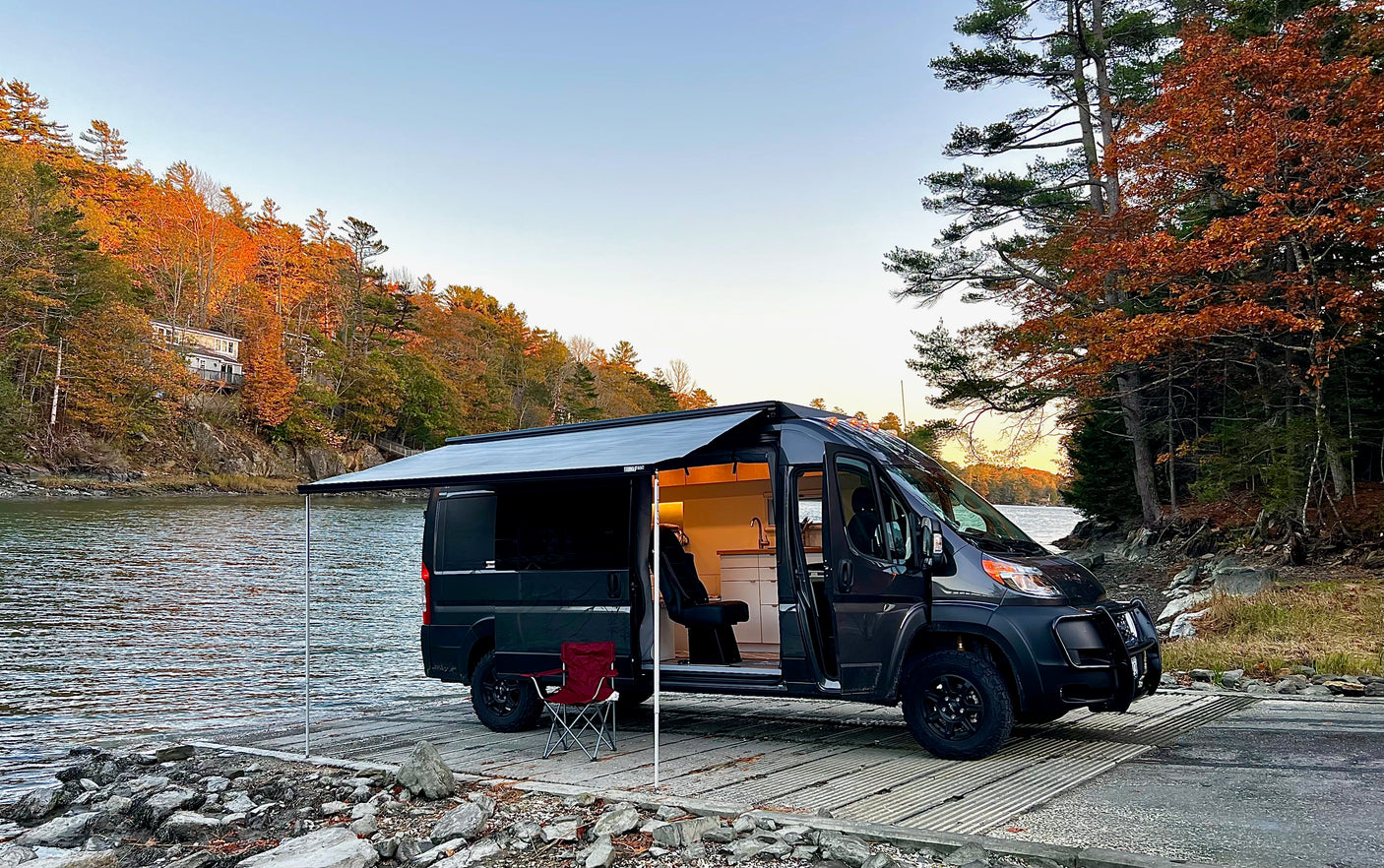 Dark gray RV rental camper van parked on a boat ramp with awning extended, overlooking autumn foliage and water. Interior visible.