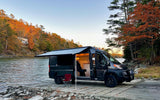 Dark gray RV rental camper van parked on a boat ramp with awning extended, overlooking autumn foliage and water. Interior visible.