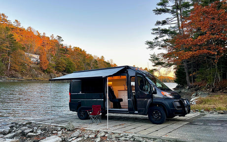 Dark gray RV rental camper van parked on a boat ramp with awning extended, overlooking autumn foliage and water. Interior visible.