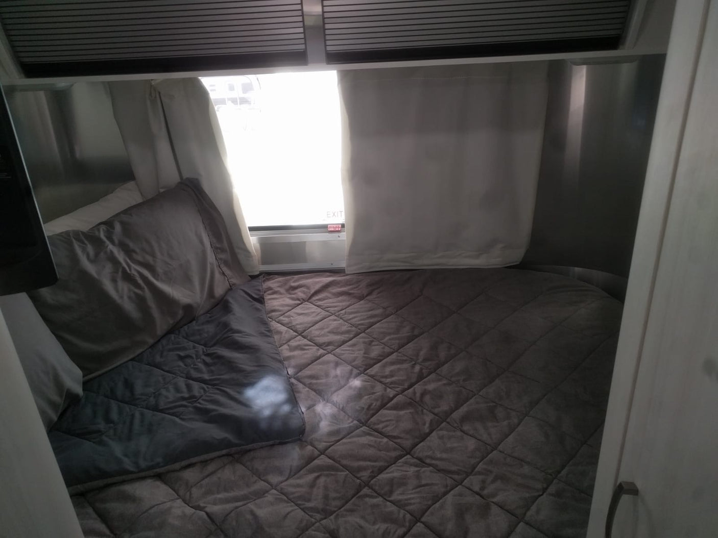 Cozy RV rental bedroom with a plush quilted bed, pillows, and a bright window featuring an "EXIT" sign.