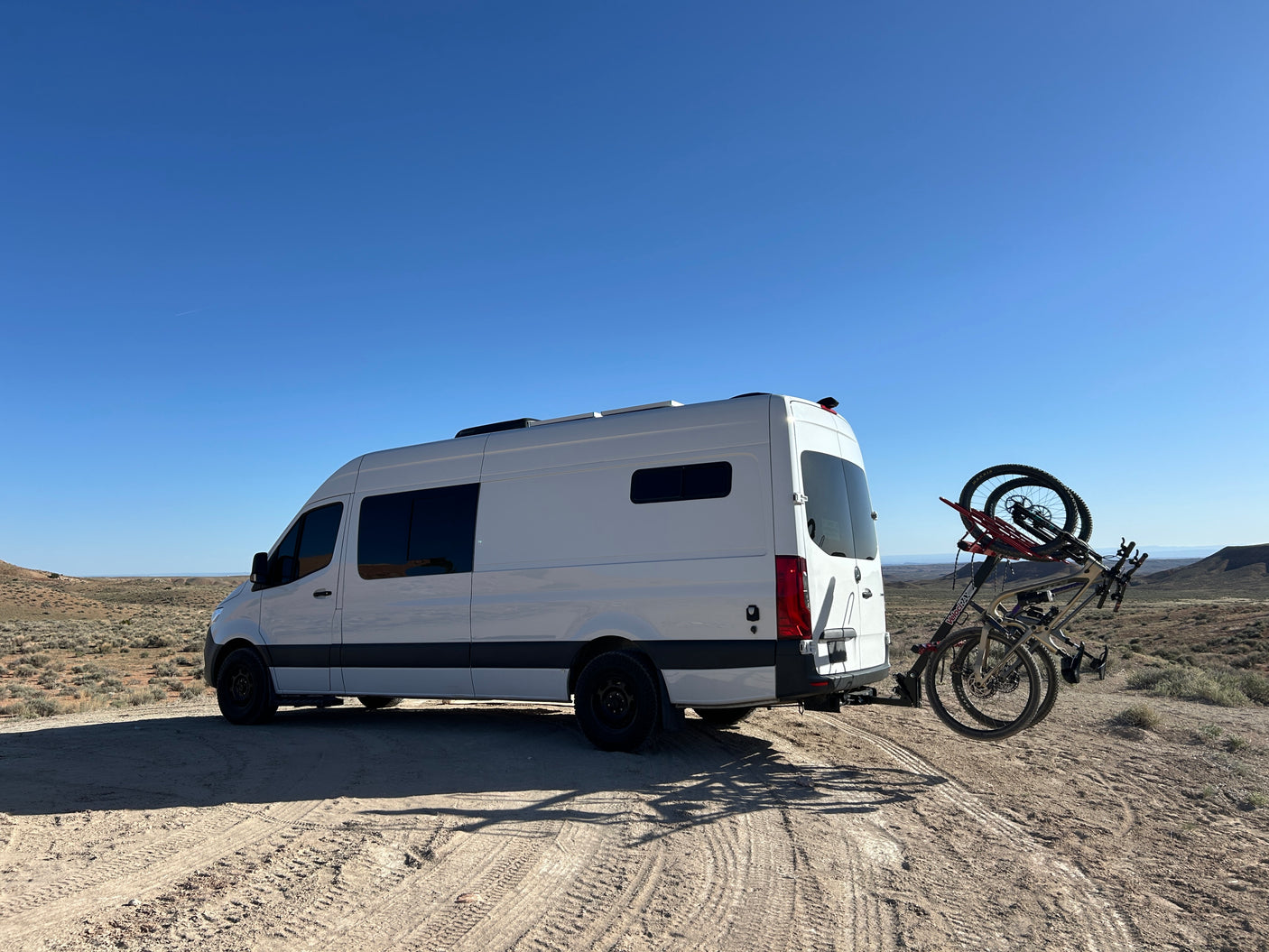 White RV rental van with mountain bikes parked on a dirt road in a sunny desert. Ready for outdoor adventures.