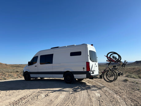 White RV rental van with mountain bikes parked on a dirt road in a sunny desert. Ready for outdoor adventures.