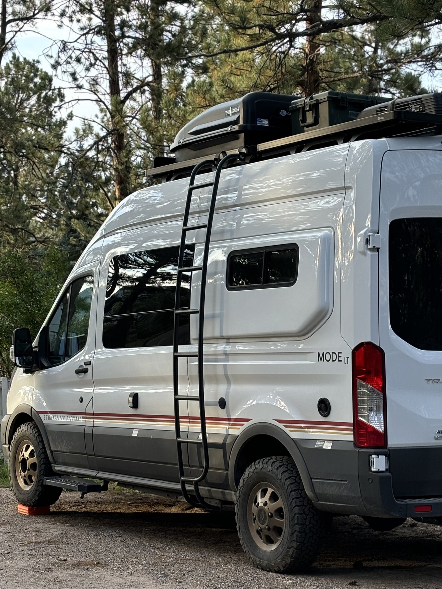 A white Storyteller Overland camper van, ideal for rv rental adventures, parked amidst pine trees, featuring a roof rack and ladder.