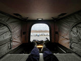 Relaxing in a pop-top RV rental. Legs extended on the bed, looking out at a large RV dealership lot with many RVs.