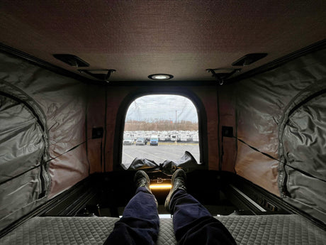 Relaxing in a pop-top RV rental. Legs extended on the bed, looking out at a large RV dealership lot with many RVs.