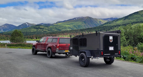 Exploring Alaska with an RV rental: Red pickup truck towing an off-road Hiker trailer, parked by a lake amidst lush mountains.