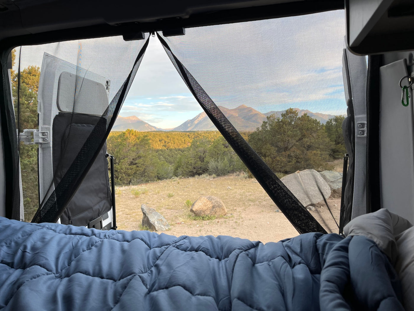  with a stunning mountain sunset. Great for remote adventures!|Cozy RV rental bed view through bug screens to majestic mountains and serene forest. Perfect for outdoor escape.|White Winnebago Solis RV rental parked on a rugged mountain road under cloudy skies — photo 2