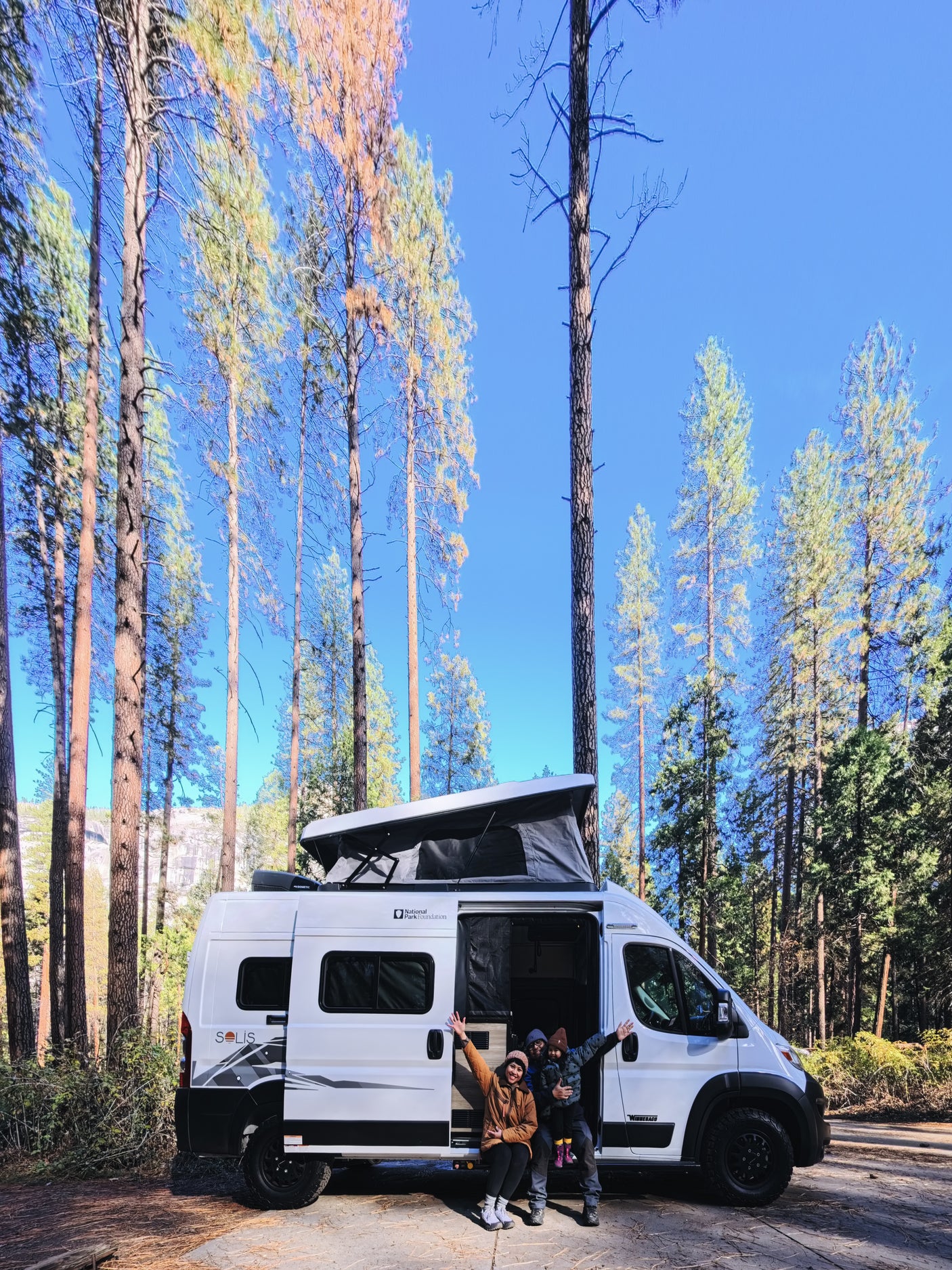 Family enjoys their white Winnebago Solis rv rental, pop-top extended, amidst tall pine trees under a blue sky, experiencing nature.