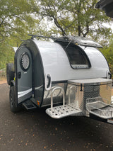 Sleek grey & white teardrop rv rental with open door, pop-up window, spare tire, and front storage box.