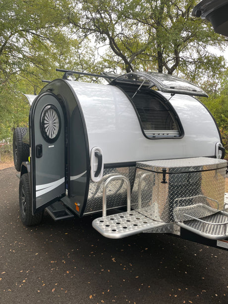 Sleek grey & white teardrop rv rental with open door, pop-up window, spare tire, and front storage box.