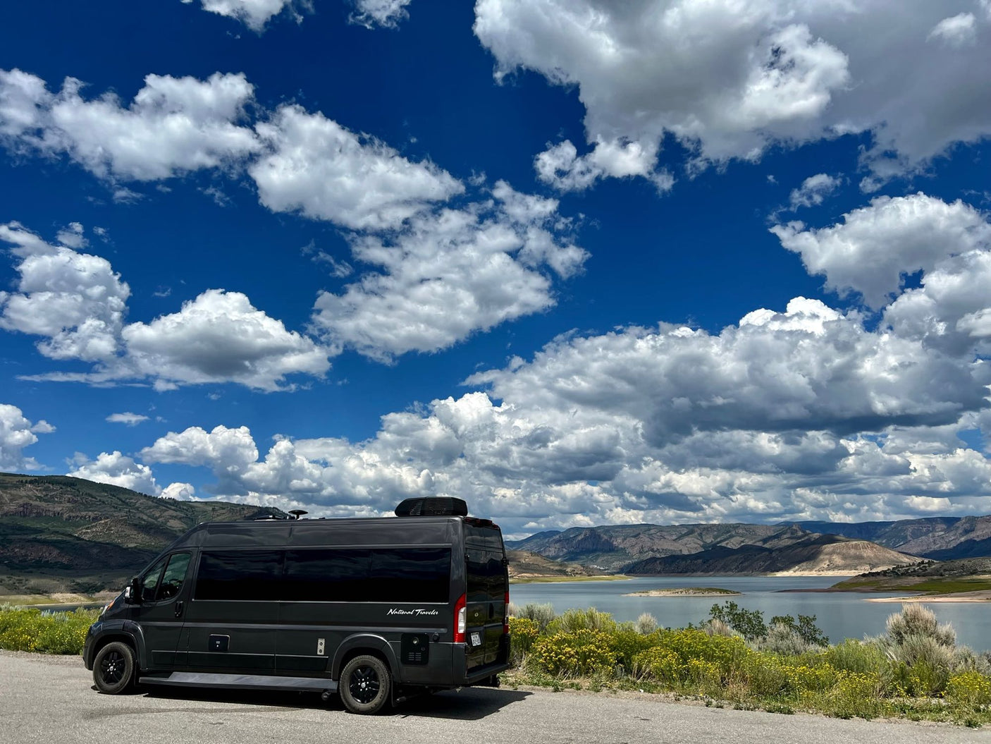 Black RV rental camper van overlooks a scenic lake with mountains under a blue, cloudy sky.