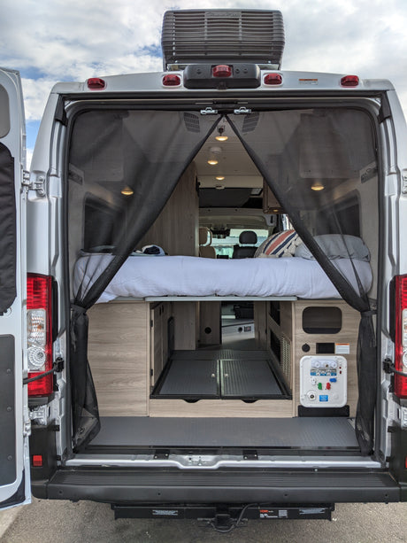 Explore this RV rental's spacious camper van interior! Features a bed, ample storage, and utility connections through open back doors.