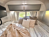 Cozy RV rental bed with striped bedding, various pillows, and a blanket, offering a relaxing view of nature.