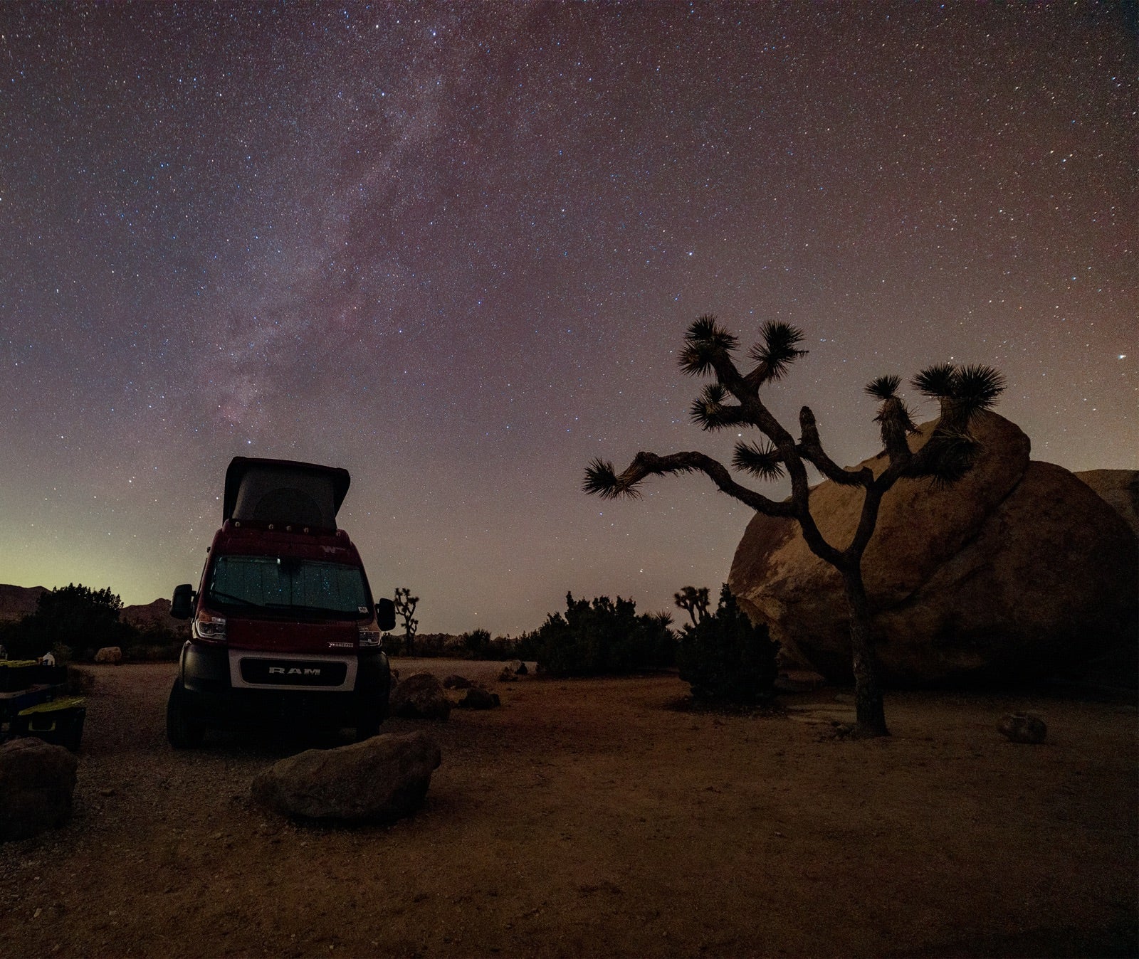 An RV rental rests under a starry night sky, featuring a Joshua tree and desert landscape. Ideal for stargazing trips.