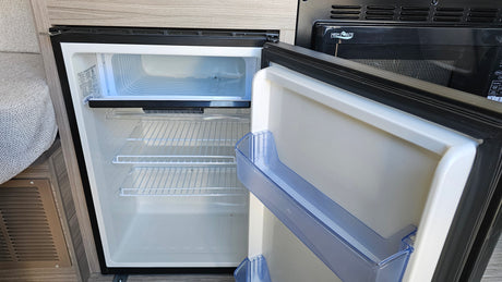 Inside an RV rental, an open compact refrigerator reveals shelves and a freezer, alongside a black microwave oven.