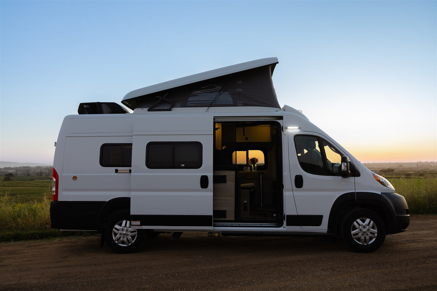 White pop-top RV rental parked on a dirt road at sunset. Its open side door reveals a bright, cozy interior, ready for adventure.