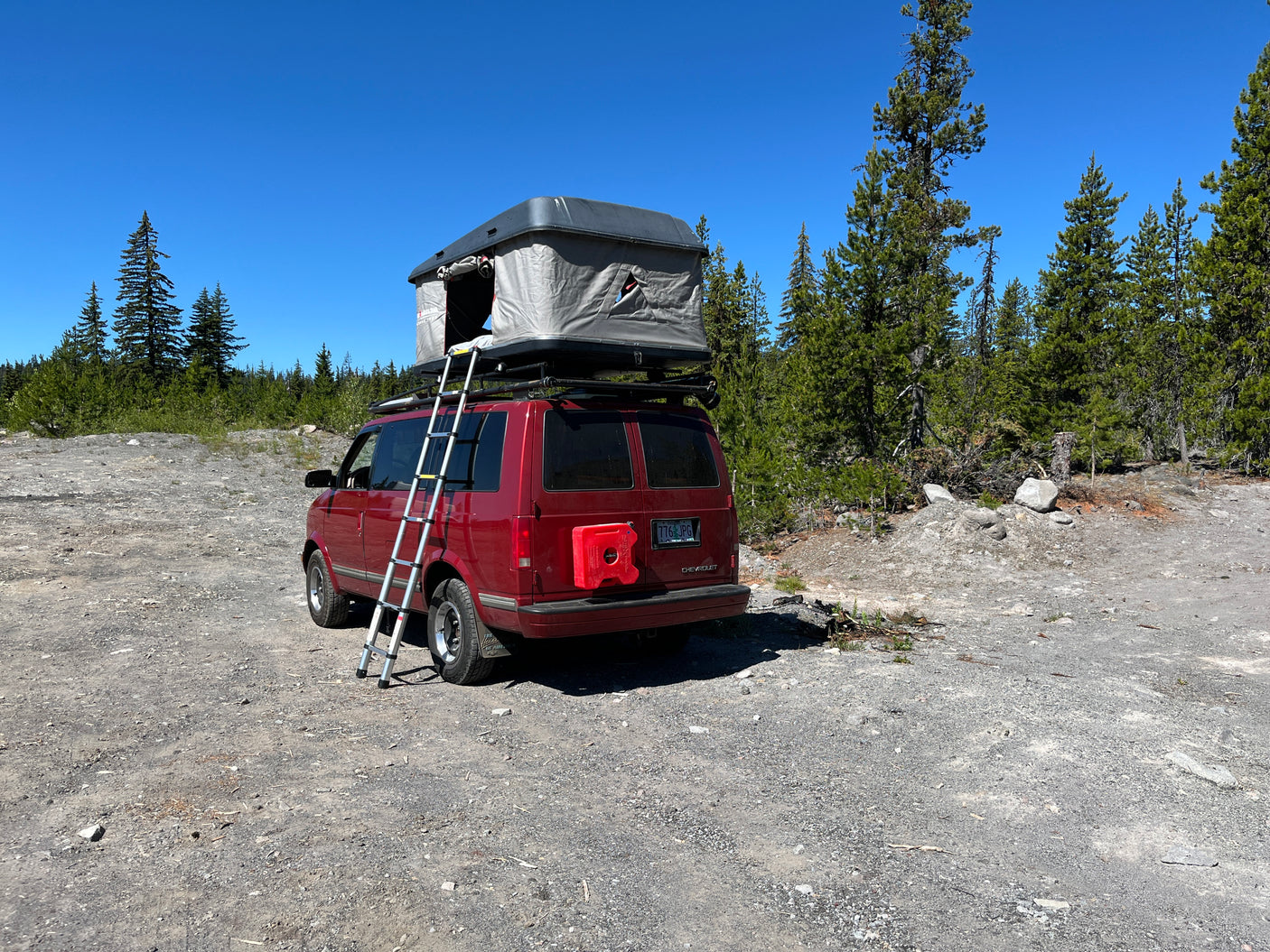 Red RV rental camper van with a rooftop tent and ladder, parked on a sunny gravel path in a forest.
