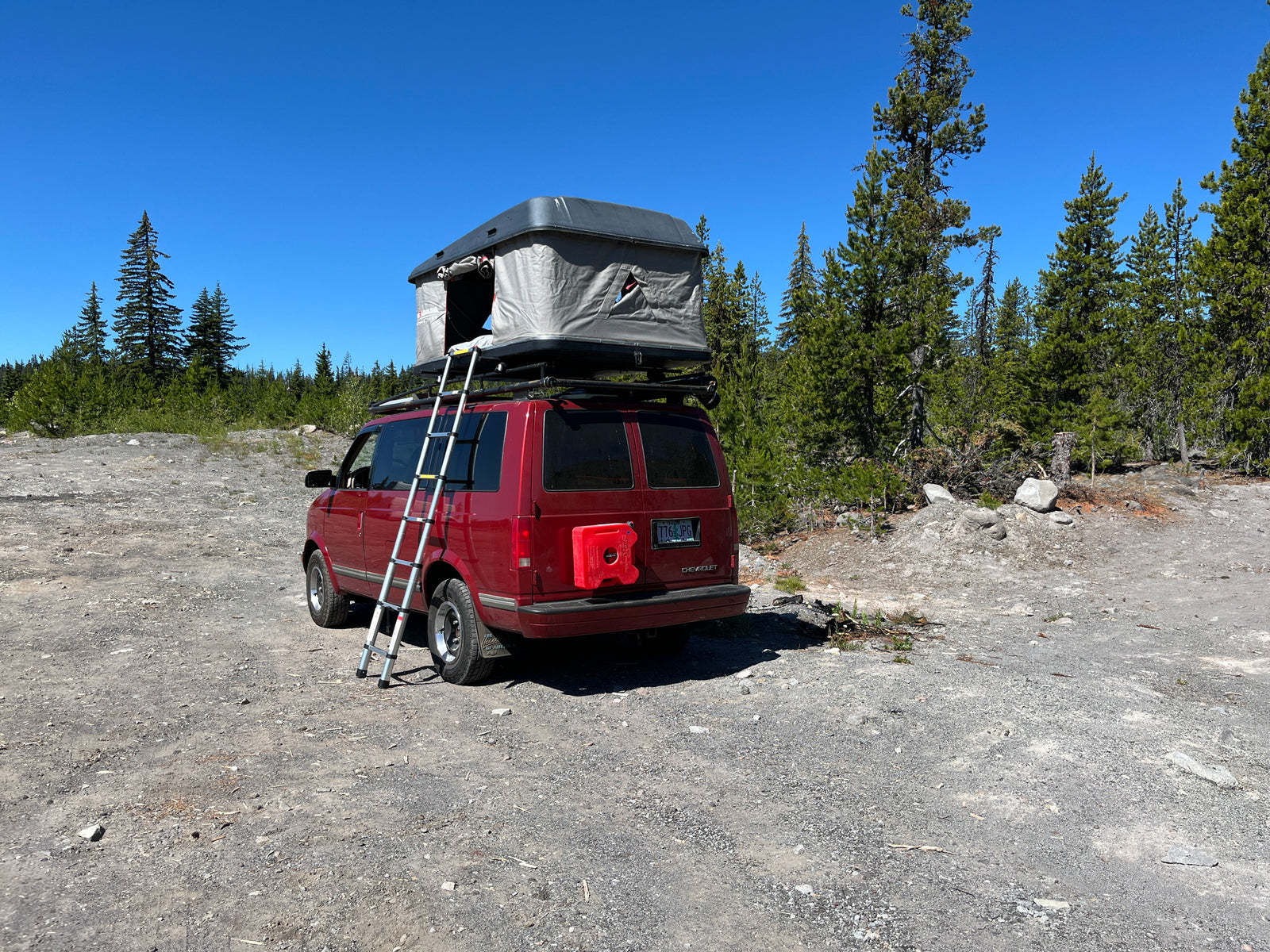 Red RV rental camper van with a rooftop tent and ladder, parked on a sunny gravel path in a forest.