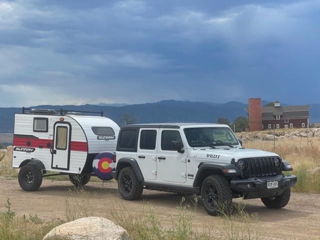 A white Jeep Wrangler tows a white SunRay RV rental, Colorado flag on spare tire. Mountains & red barn in distance.