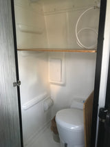 Compact RV rental wet bath with white walls, toilet, wooden shelf, and coiled shower hose. Efficient use of space.