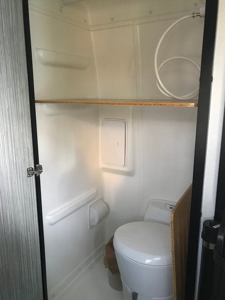 Compact RV rental wet bath with white walls, toilet, wooden shelf, and coiled shower hose. Efficient use of space.