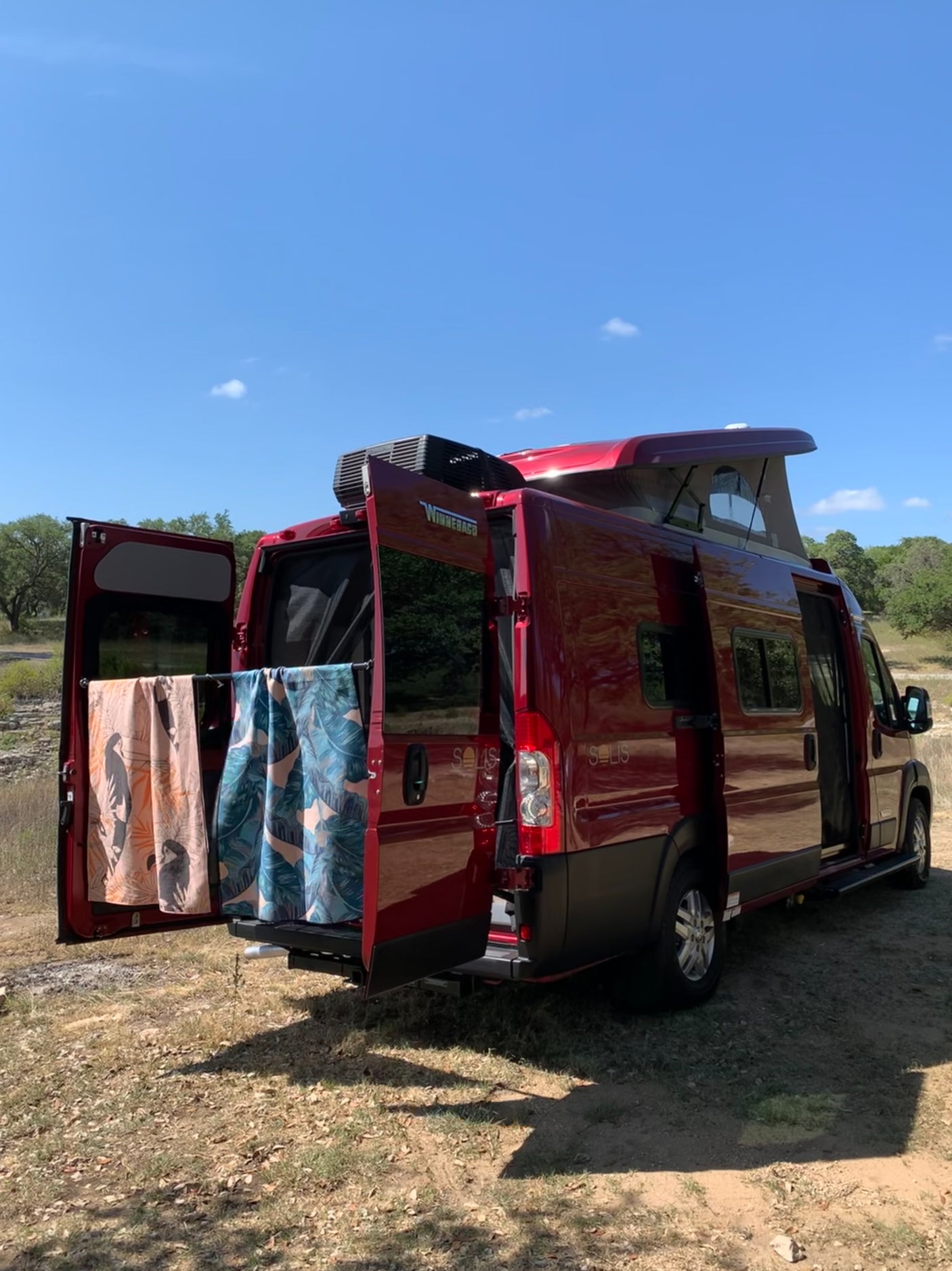 Red Winnebago Solis RV rental parked. Pop-top up, rear doors open with towels. Perfect for outdoor adventures!