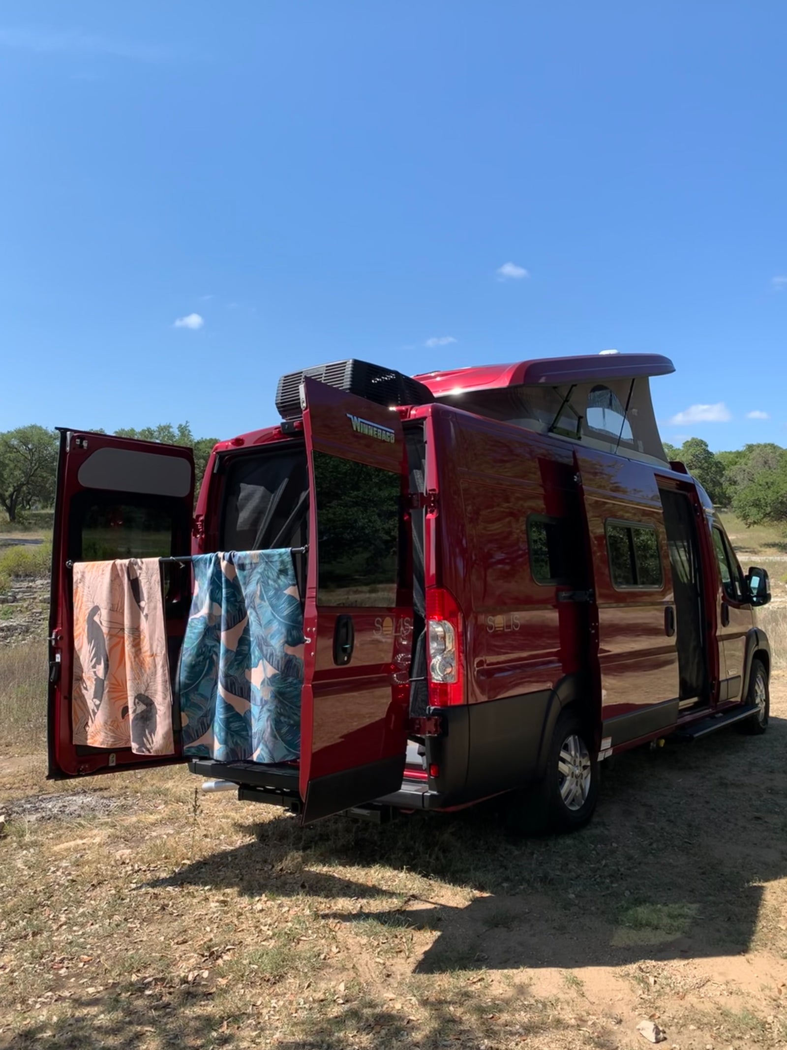 Red Winnebago Solis RV rental parked. Pop-top up, rear doors open with towels. Perfect for outdoor adventures!