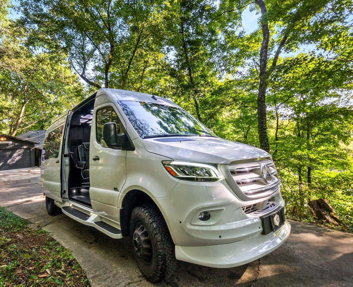 White Mercedes-Benz Sprinter camper van, an ideal RV rental, parked with its side door open in a sunny, wooded drive.