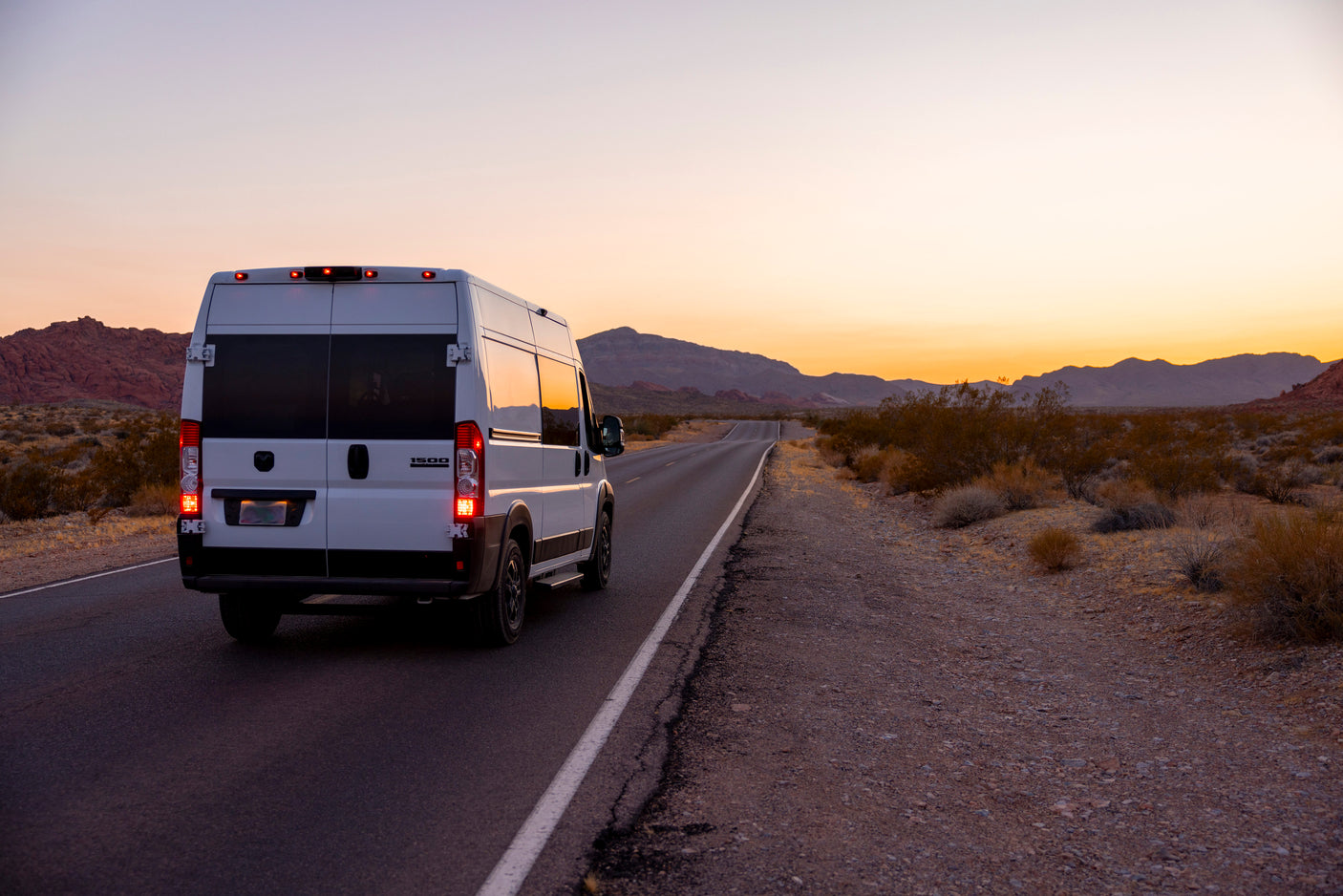 White RV rental van on a desert road at sunset, framed by mountains. Your ideal vehicle for scenic road trips & adventures.
