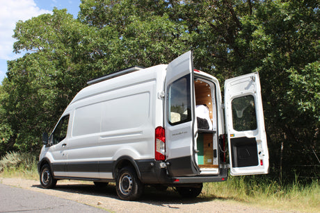 This white Ford Transit camper van, ideal for RV rental, is parked outdoors with its rear doors open, revealing a cozy wooden interior.