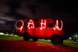 Light-painted 'OAHU' glows red over an RV rental on a grassy field at night, with stars and clouds visible above.