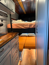 RV rental interior with light grey cabinets, wood counters, cozy upper bed, and lower orange cushion seating.