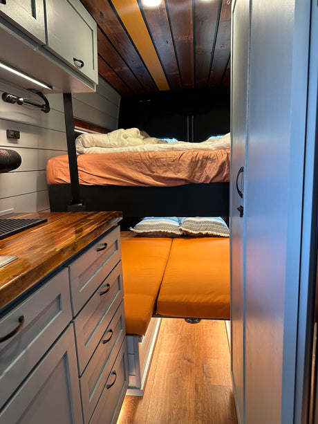 RV rental interior with light grey cabinets, wood counters, cozy upper bed, and lower orange cushion seating.