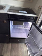 RV rental kitchenette featuring a sleek countertop, integrated stovetop, sink, and an open, empty mini-fridge below.