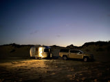 Lighted RV rental and truck parked for beach camping on sand dunes under a starry twilight sky.