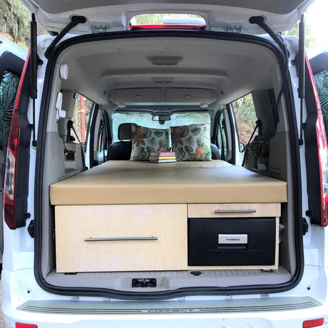 Inside a white RV rental van, a custom bed, storage, & fridge create a cozy camper setup for travel.