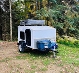 Compact teardrop camper, perfect for an RV rental adventure, parked in an autumn forest with a rooftop tent and cooler.