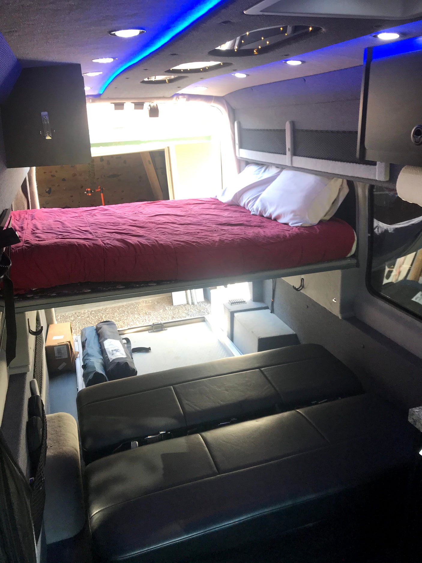 Interior view of an rv rental camper van, featuring a bed with red bedding, black passenger seats, and rear storage. Blue LED lighting.