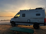 RV rental adventure! Gray camper van by a lake at golden hour, a green picnic table in front. Perfect travel & camping scene.