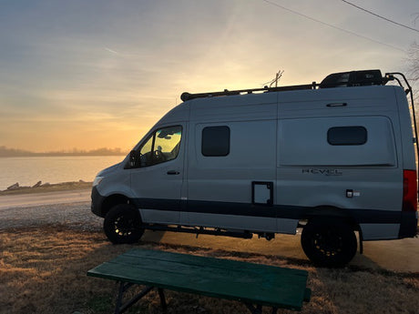 RV rental adventure! Gray camper van by a lake at golden hour, a green picnic table in front. Perfect travel & camping scene.