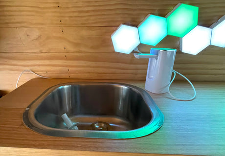 Compact RV rental utility nook featuring a small sink, automatic dispenser, and glowing hexagonal lights on wooden walls.