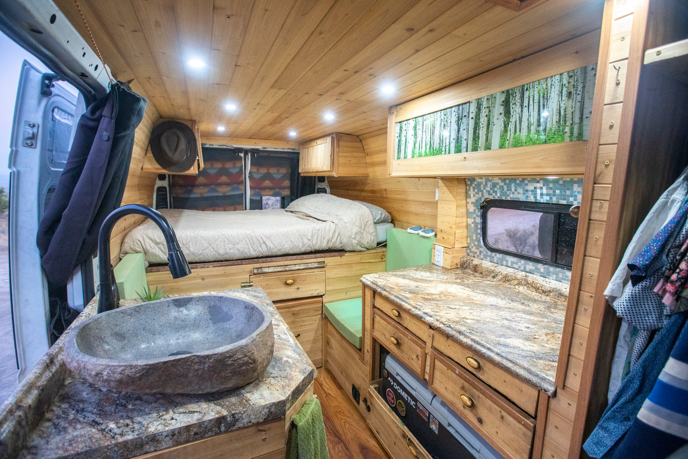 Cozy RV rental interior with custom wood, a bed, stone sink, and granite counter kitchen. Perfect camper van experience.